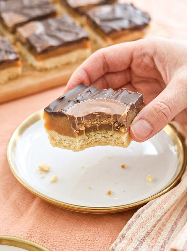 A hand holds a chocolate caramel bar with a bite taken out, showing layers of shortbread, caramel, and a peanut butter cup on top.