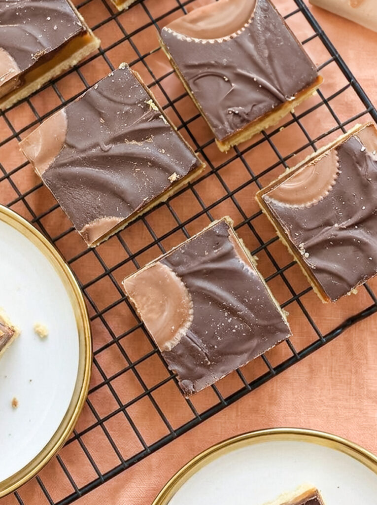 Square millionaire bars with layers of shortbread, caramel, and chocolate topped with peanut butter cups on a wire rack.