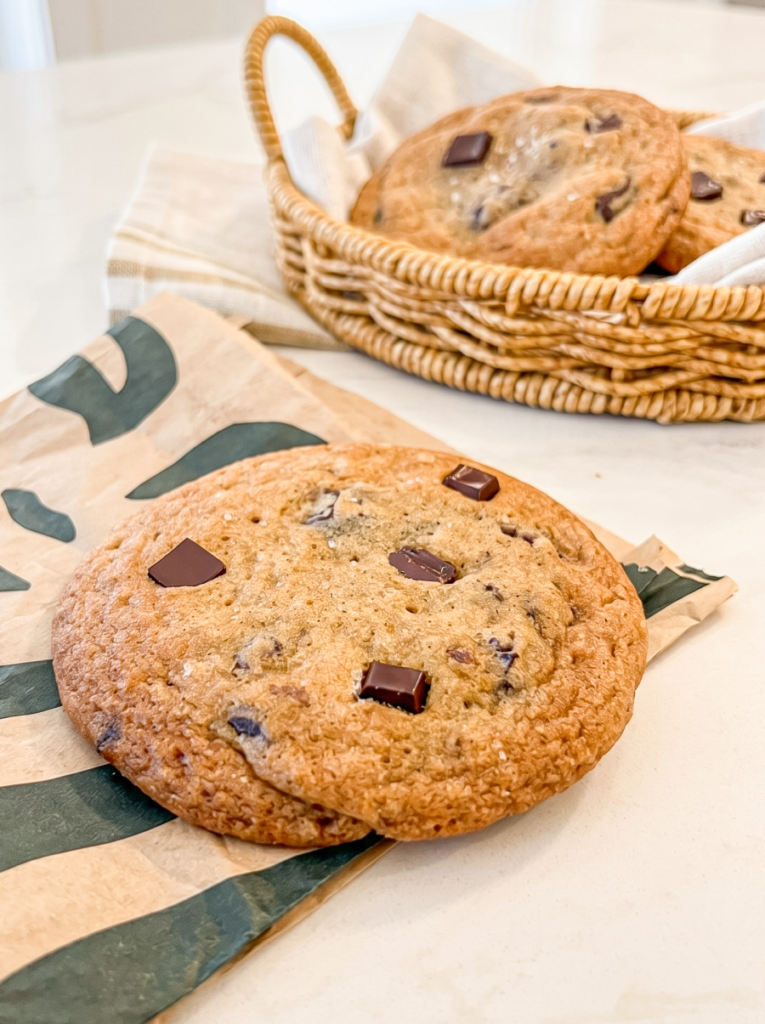 A large chocolate chunk cookie with sea salt rests on a brown paper bag, with a wicker basket of more cookies in the background.