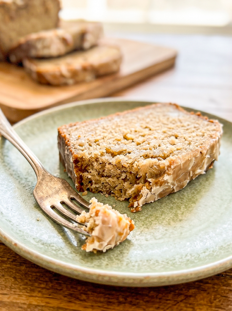 A thick slice of glazed banana bread with a moist, golden crumb on a sage green ceramic plate with a copper fork and bite taken. Sliced loaf visible on wooden board in background.
