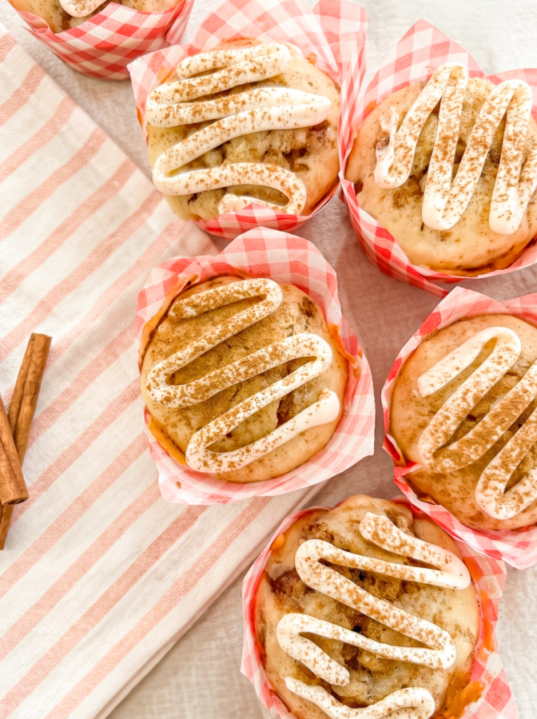Six cinnamon roll muffins with white icing and cinnamon sprinkles in gingham liners, next to a striped towel and cinnamon sticks.