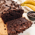 A rich double chocolate banana bread loaf with a slice cut, topped with chocolate chips, next to ripe bananas and a bowl of chips.