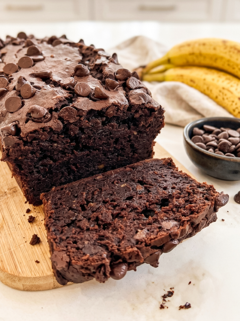 A rich double chocolate banana bread loaf with a slice cut, topped with chocolate chips, next to ripe bananas and a bowl of chips.