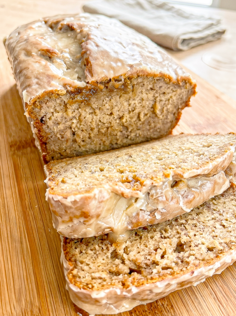 Sliced banana bread loaf with espresso glaze dripping down the sides, displayed on a wooden cutting board with a striped kitchen towel in the background. The moist interior crumb is visible in the sliced portions.