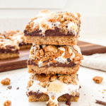 A stack of three biscoff s'mores bars with gooey melted chocolate and toasted marshmallows, crumbs scattered on a white counter.
