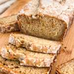 Sliced banana bread loaf with glossy espresso glaze dripping down the sides, showing moist interior studded with banana pieces. Multiple slices arranged on a wooden cutting board with a white kitchen towel in the background.