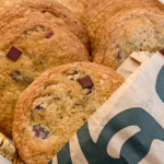 Golden-brown chocolate chunk cookies are piled in a basket, with one peeking out of a brown paper Starbucks bag.