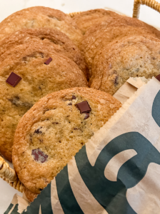 Golden-brown chocolate chunk cookies are piled in a basket, with one peeking out of a brown paper Starbucks bag.