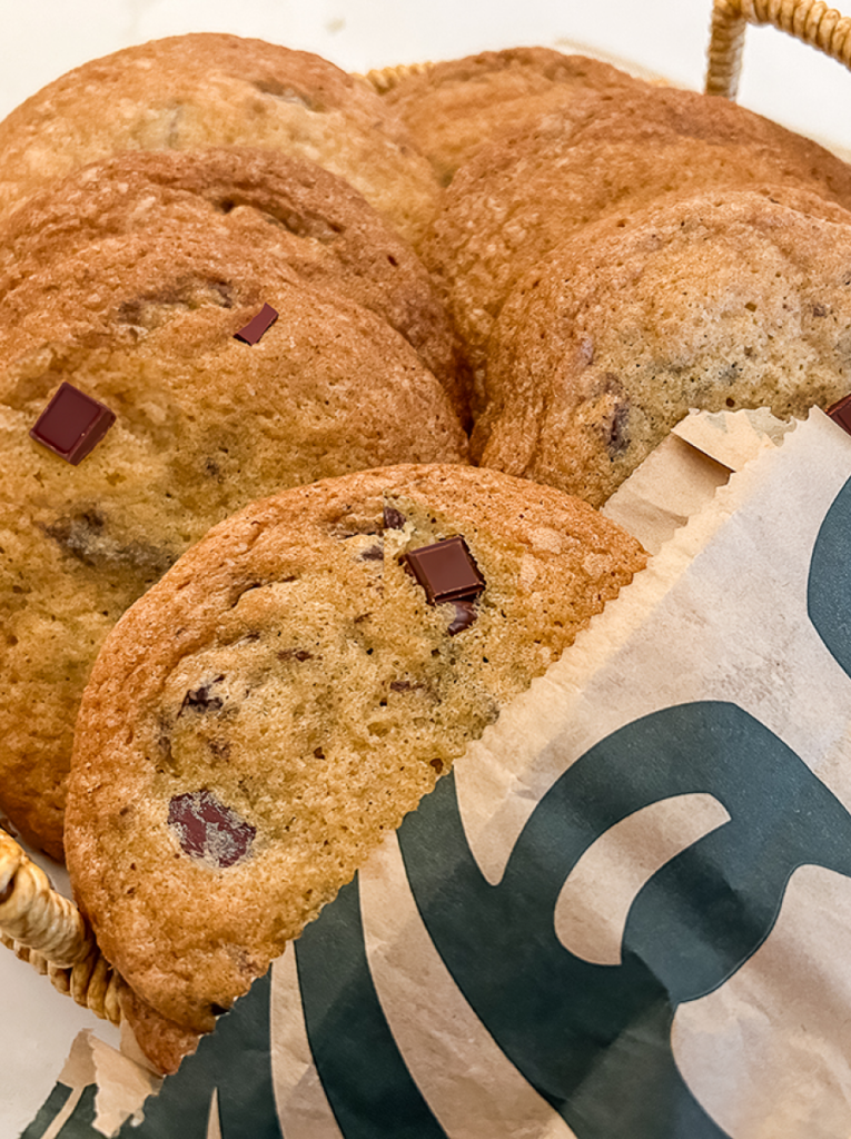 Golden-brown chocolate chunk cookies are piled in a basket, with one peeking out of a brown paper Starbucks bag.