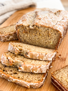 Sliced banana bread loaf with glossy espresso glaze dripping down the sides, showing moist interior studded with banana pieces. Multiple slices arranged on a wooden cutting board with a white kitchen towel in the background.