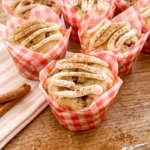 Muffins in red checkered wrappers topped with white icing and cinnamon sit on a wood table with cinnamon sticks and a whisk.
