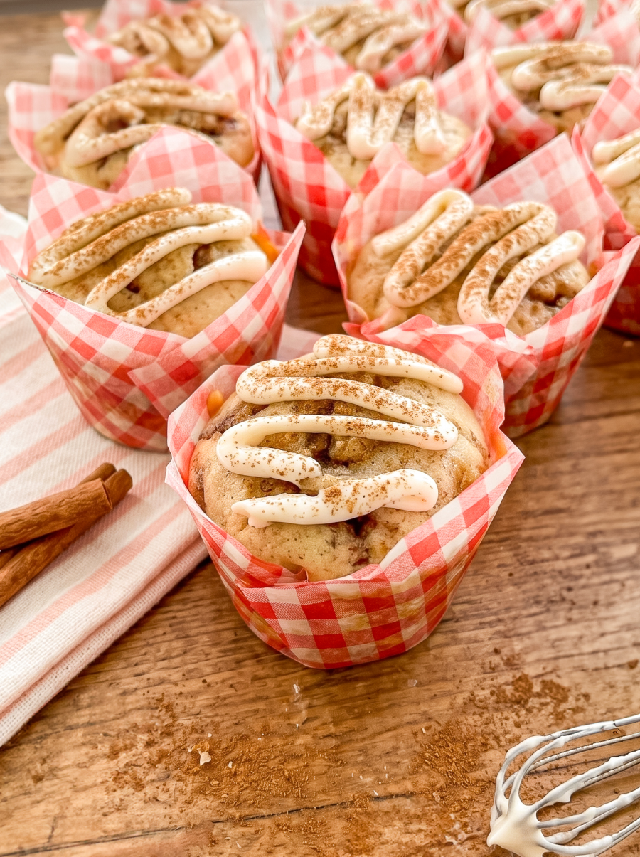 Muffins in red checkered wrappers topped with white icing and cinnamon sit on a wood table with cinnamon sticks and a whisk.