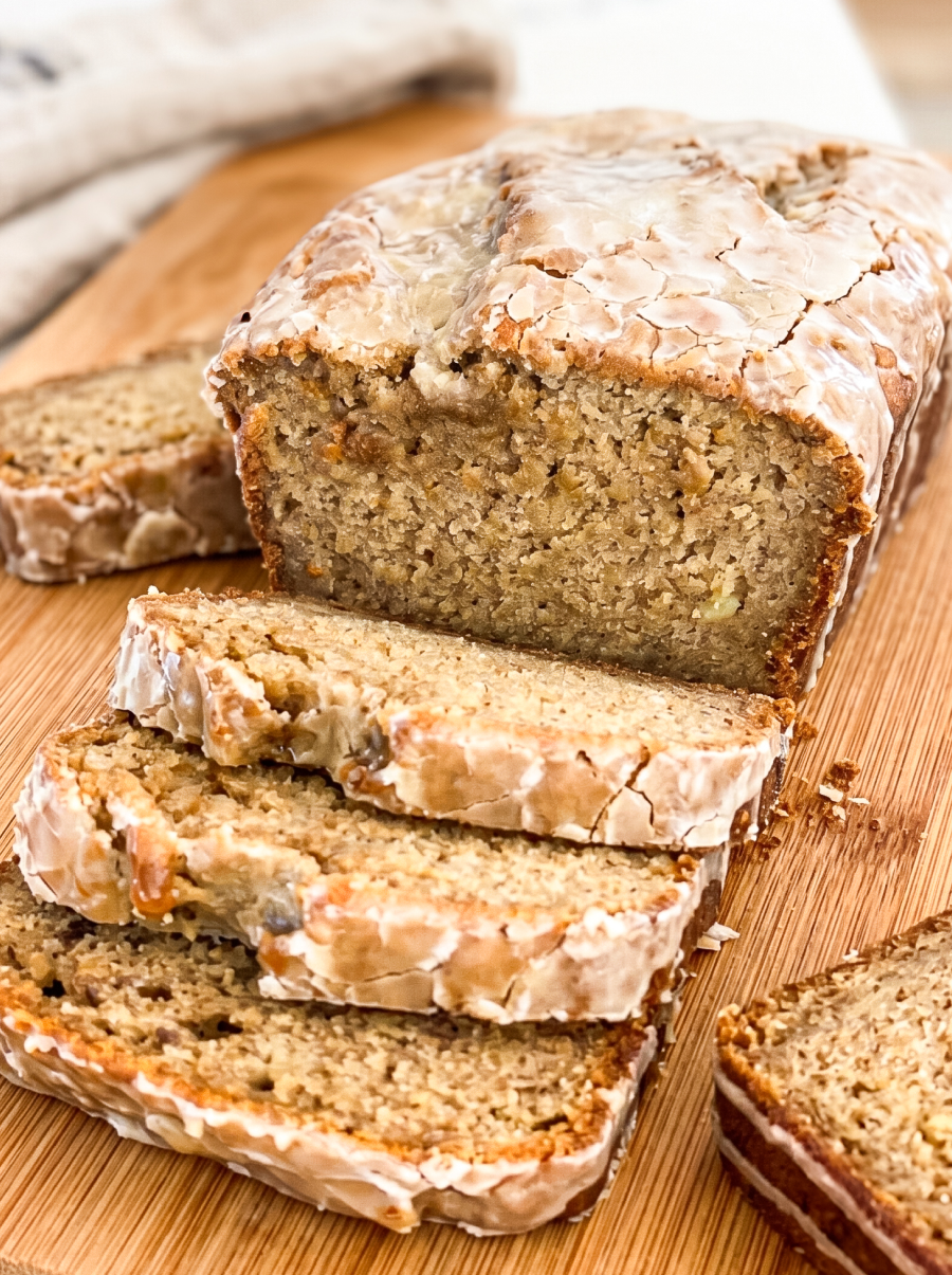 Sliced banana bread loaf with glossy espresso glaze dripping down the sides, showing moist interior studded with banana pieces. Multiple slices arranged on a wooden cutting board with a white kitchen towel in the background.