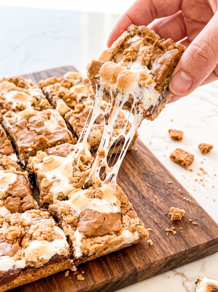 A hand lifts a biscoff s'mores bar from a wooden board, showcasing its gooey, stretchy melted marshmallow and crumbly topping.