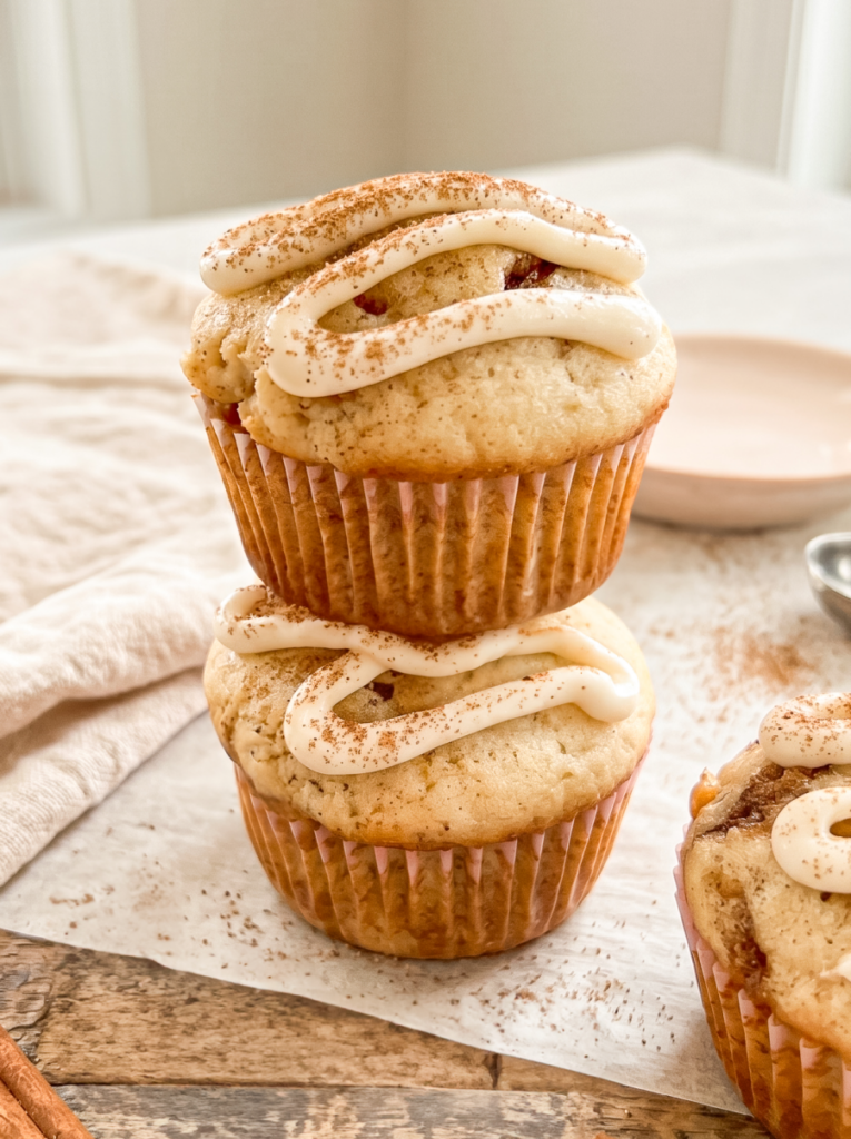 Two cinnamon roll muffins, topped with white icing and cinnamon, are stacked on parchment paper,
