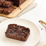 A slice of rich double chocolate banana bread with chocolate chips rests on a white plate, with the full loaf and a ripe banana visible.