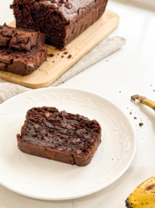 A slice of rich double chocolate banana bread with chocolate chips rests on a white plate, with the full loaf and a ripe banana visible.