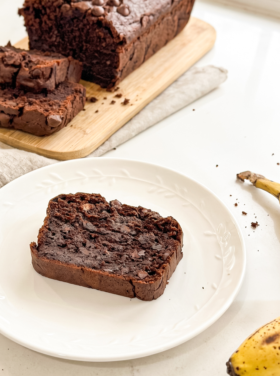 A slice of rich double chocolate banana bread with chocolate chips rests on a white plate, with the full loaf and a ripe banana visible.