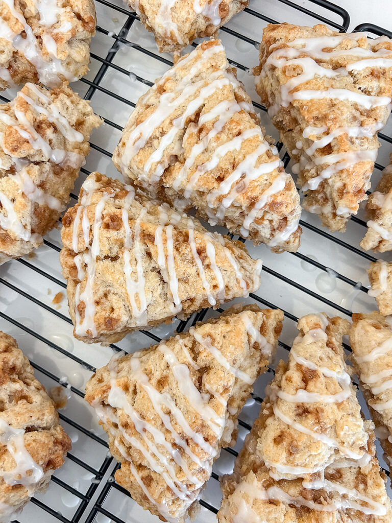 Multiple triangular cinnamon toffee scones with a white glaze drizzle rest on a black cooling rack.