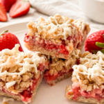 A stack of oat bars with a bright red strawberry filling and white drizzle sits on a white plate, surrounded by fresh strawberries.