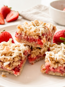 A stack of oat bars with a bright red strawberry filling and white drizzle sits on a white plate, surrounded by fresh strawberries.