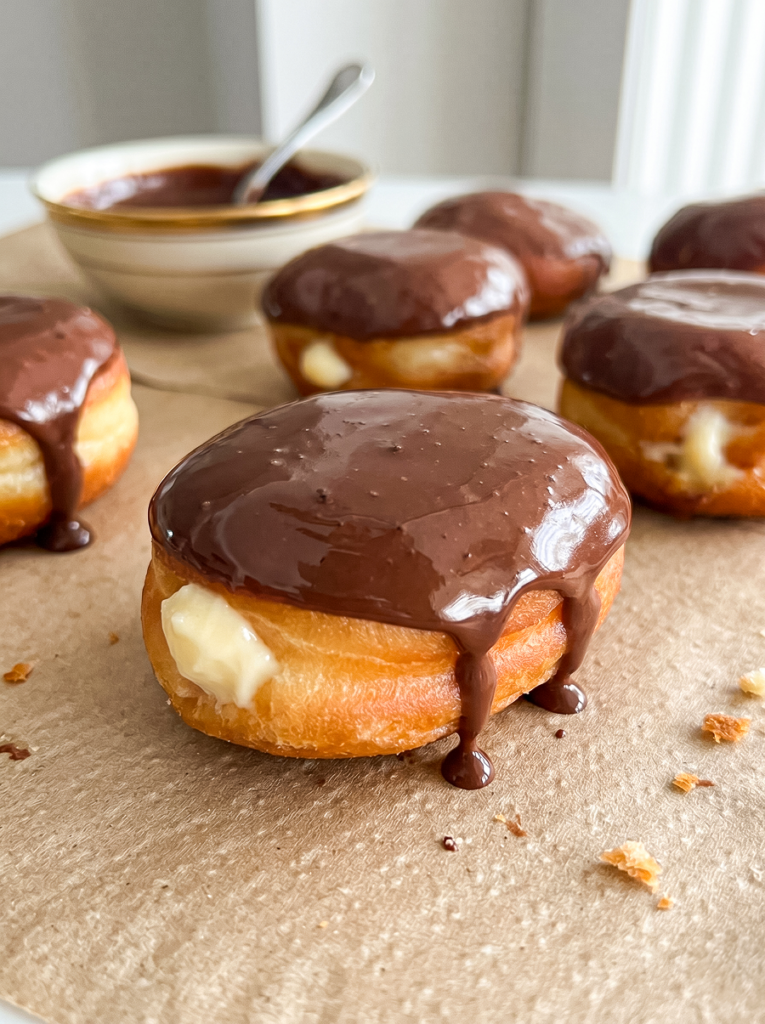Golden Boston cream donuts with creamy filling and glossy chocolate glaze dripping down on parchment paper, with a bowl of glaze in the background.