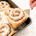 A hand uses a spatula to lift a warm, iced banana pecan cinnamon roll from a baking pan filled with more rolls, ready to serve.