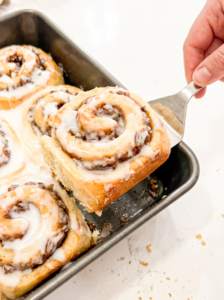 A hand uses a spatula to lift a warm, iced banana pecan cinnamon roll from a baking pan filled with more rolls, ready to serve.