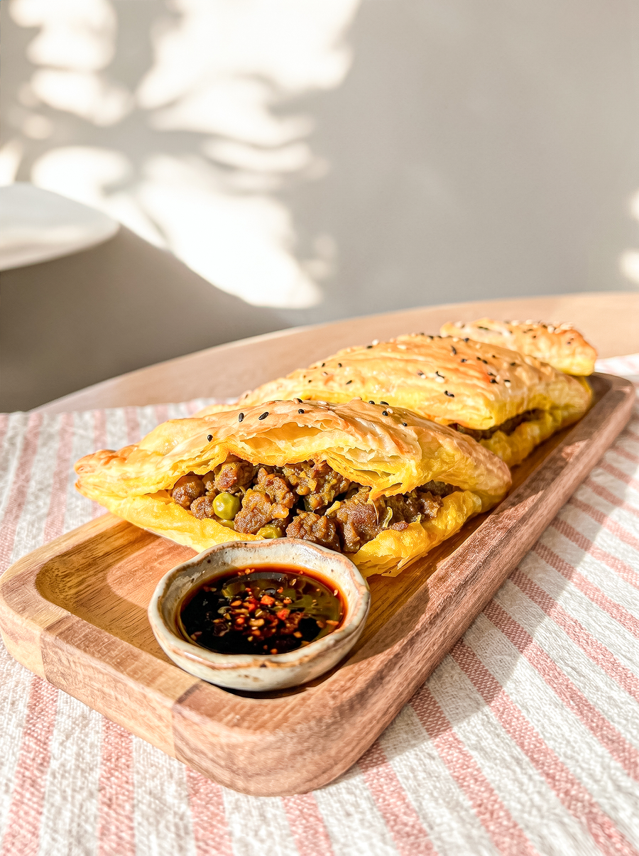 Three golden, flaky pastry puffs filled with savory beef and peas, served on a wooden tray with a chili dipping sauce.
