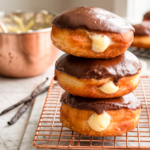 Three Boston cream donuts with chocolate glaze and vanilla cream filling are stacked on a copper rack, with vanilla beans and chocolate curls nearby.