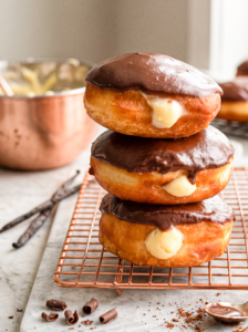 Three Boston cream donuts with chocolate glaze and vanilla cream filling are stacked on a copper rack, with vanilla beans and chocolate curls nearby.