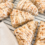 Several triangular cinnamon toffee scones with white icing drizzle are cooling on a black wire rack, with a linen napkin nearby.