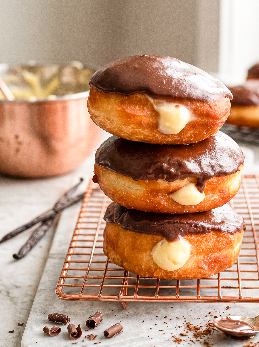 Three Boston cream donuts with chocolate glaze and vanilla cream filling are stacked on a copper rack, with vanilla beans and chocolate curls nearby.