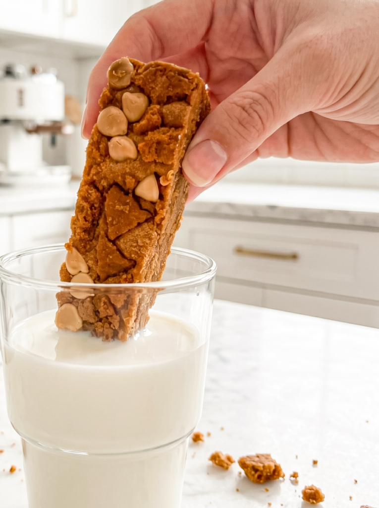 A hand dips a Biscoff cookie butter bar, topped with light chips and crumbs, into a glass of milk in a bright kitchen.