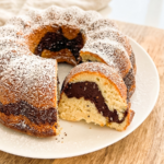 A marble bundt cake, dusted with powdered sugar, sits on a white plate with a slice removed, revealing the chocolate and vanilla swirl.