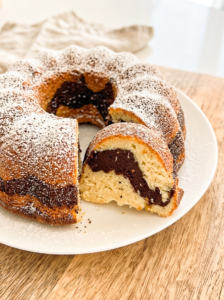 A marble bundt cake, dusted with powdered sugar, sits on a white plate with a slice removed, revealing the chocolate and vanilla swirl.