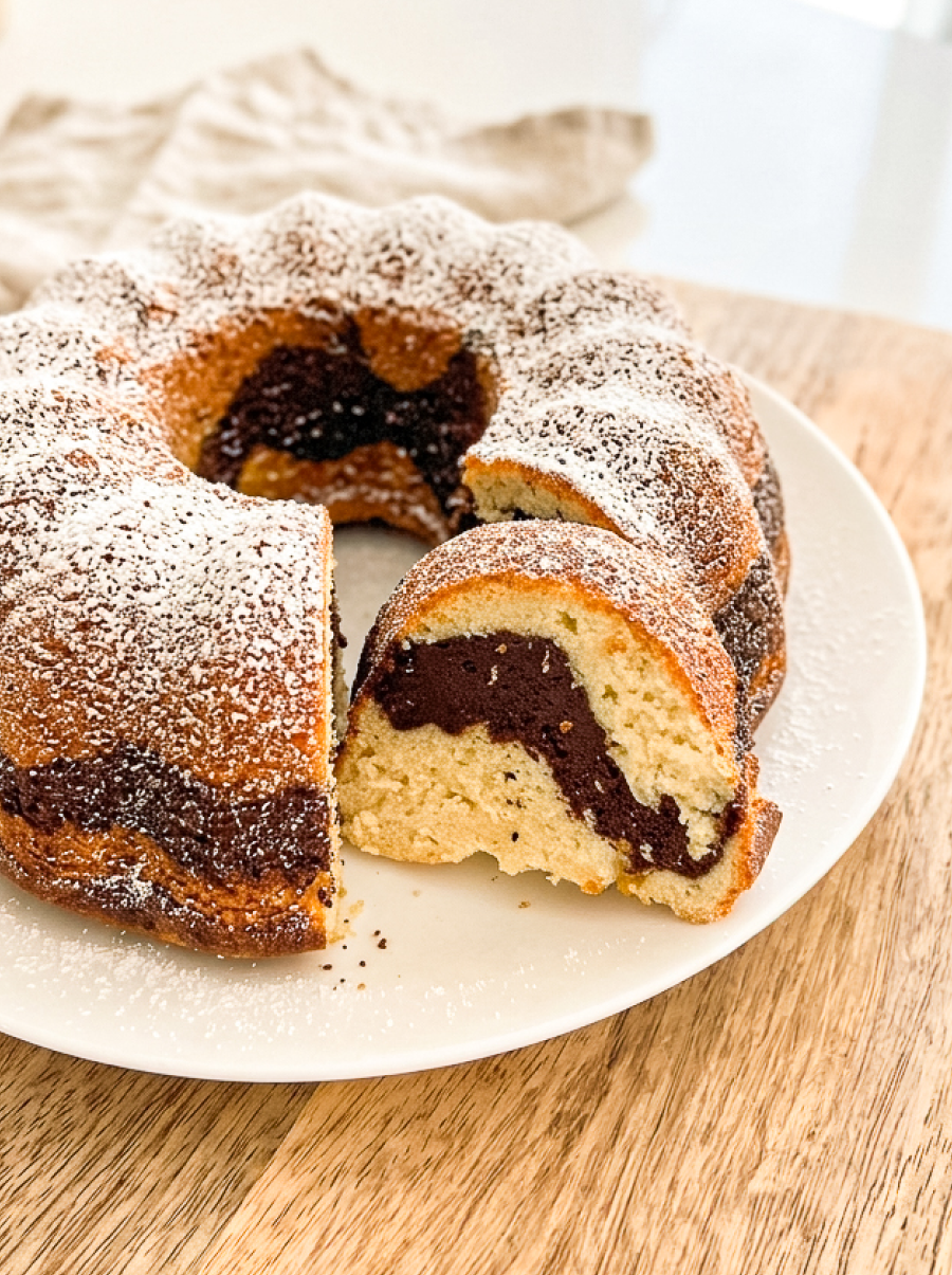 A marble bundt cake, dusted with powdered sugar, sits on a white plate with a slice removed, revealing the chocolate and vanilla swirl.