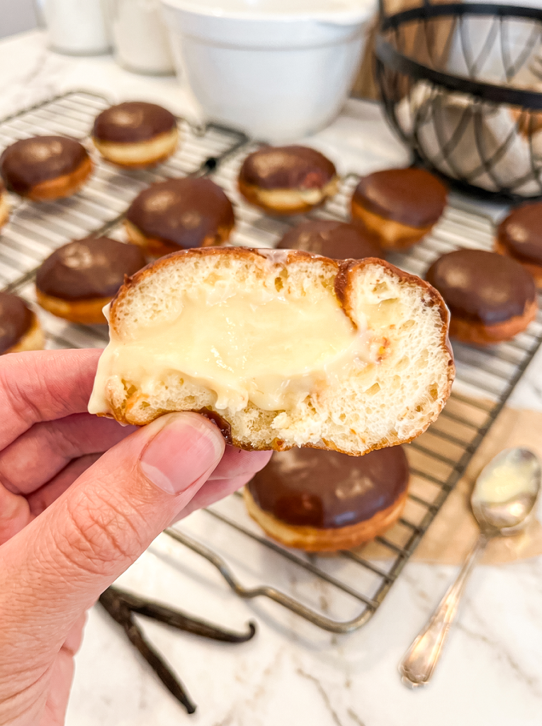 A hand holds a Boston cream donut cut in half, revealing its creamy vanilla filling, with chocolate-glazed donuts on a cooling rack.