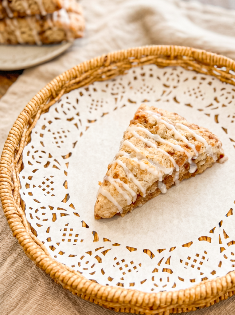 A triangular cinnamon toffee scone with white icing drizzle sits on a paper doily in a wicker basket, with another scone blurred in the background.