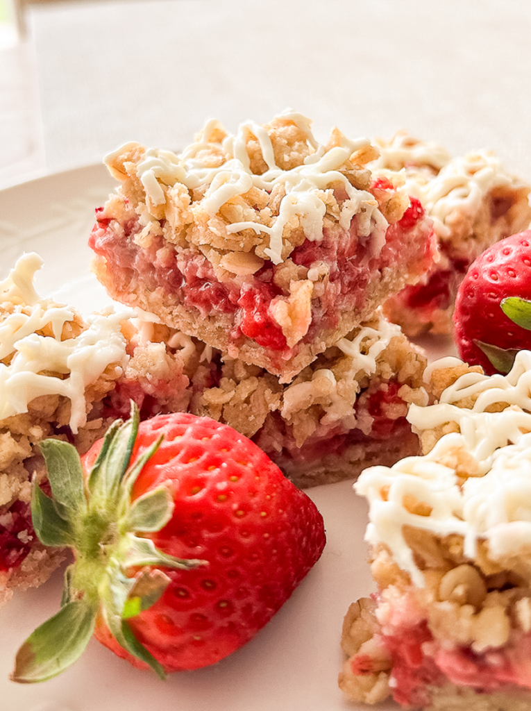 A close-up of several square oat bars with a strawberry filling and white drizzle, arranged on a white plate with fresh strawberries.
