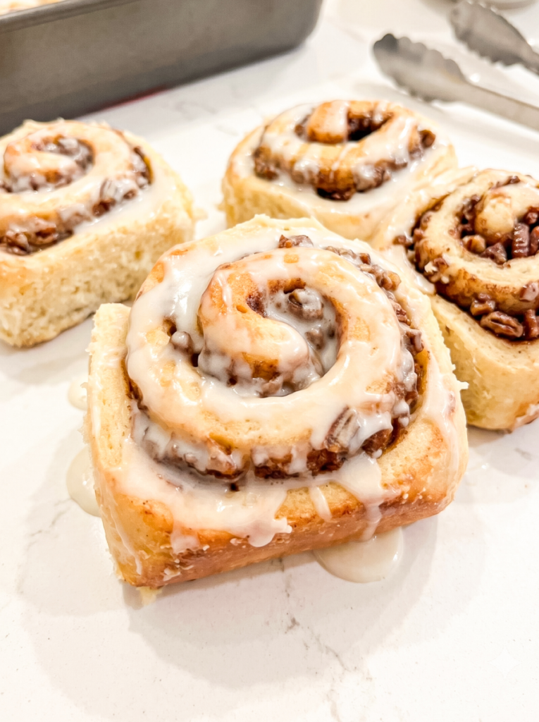 Four banana pecan cinnamon rolls covered in white icing sit on a counter, with a baking pan and tongs in the background.