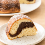 A slice of bundt cake with a chocolate swirl and powdered sugar on top sits on a speckled plate, with more cake in the background.
