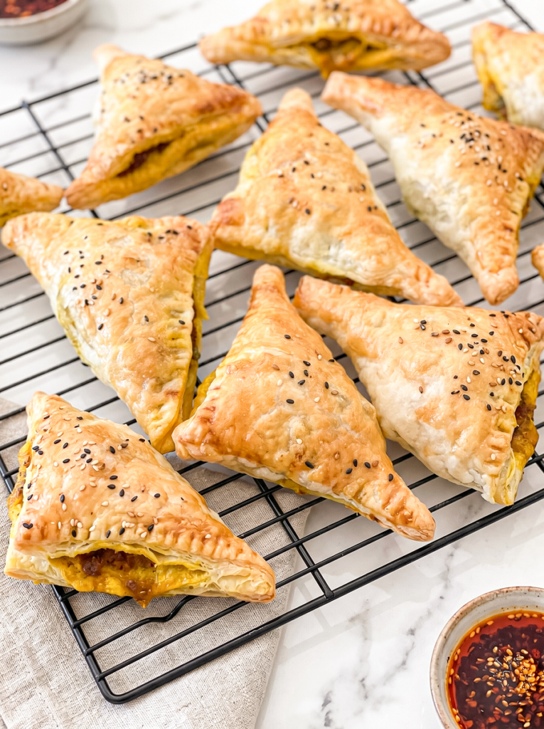 Golden-brown triangular curry beef puffs with sesame seeds are cooling on a black wire rack, with a small bowl of chili oil nearby.