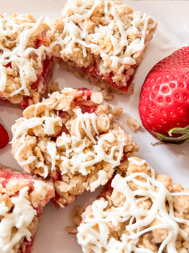 Several oat crumble bars with strawberry filling and white icing drizzle are arranged on a white plate next to a whole fresh strawberry.