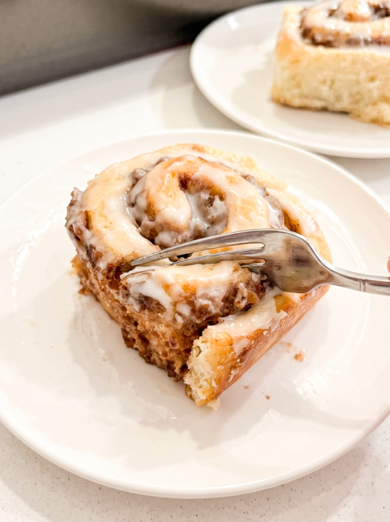 A fork takes a bite from a sweet, iced banana pecan cinnamon roll on a white plate, with another roll in the background.