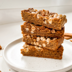 A stack of four golden brown biscoff cookie butter bars, topped with white chips and crumbs, sits on a white plate in a bright kitchen.