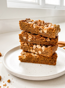 A stack of four golden brown biscoff cookie butter bars, topped with white chips and crumbs, sits on a white plate in a bright kitchen.