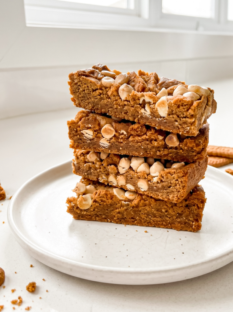 A stack of four golden brown biscoff cookie butter bars, topped with white chips and crumbs, sits on a white plate in a bright kitchen.