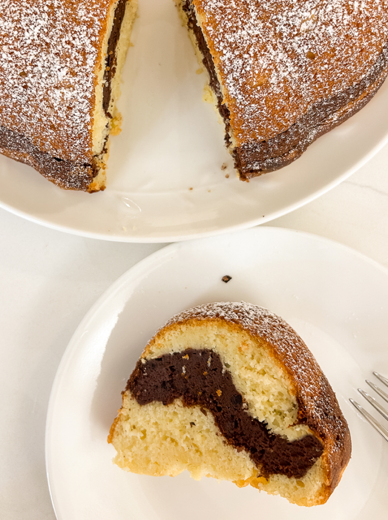 A marble bundt cake, dusted with powdered sugar, on a white plate, with one slice served on another plate nearby.
