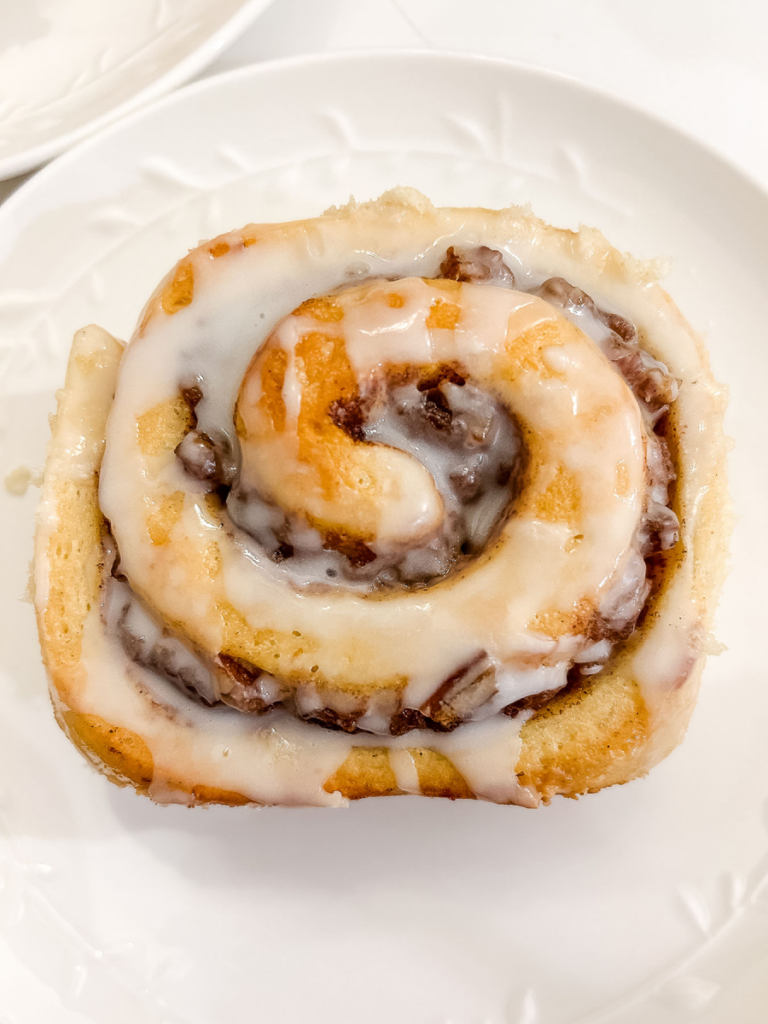 A single, glazed banana pecan cinnamon roll with a visible spiral of filling sits on a white plate.